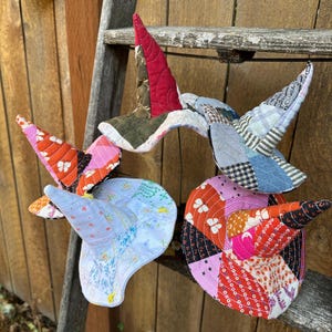 May include: A collection of handmade, whimsical fabric sculptures in the shape of birds. Each bird features a unique patchwork design with vibrant colors and patterns, including reds, oranges, pinks, blues, and greens. The birds are displayed on a rustic wooden ladder.