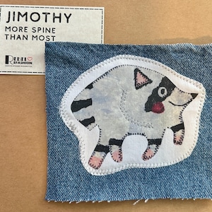 May include: A blue denim fabric patch featuring a white, black, and pink embroidered raccoon-like creature with a striped tail. A rectangular card is included with the text: JIMOTHY MORE SPINE THAN MOST, REBEL REFASHION MADE IN SPOKANE WASHINGTON.