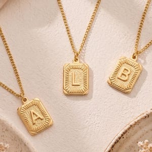 May include: Three gold-colored initial necklaces with rectangular pendants. The pendants feature the letters A, L, and B, each with a textured border and sunburst design. The necklaces are displayed on a neutral surface.