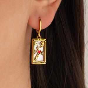 May include: Gold hoop earrings with a rectangular charm featuring a white tarot card with a red heart and the text "THE LOVERS".