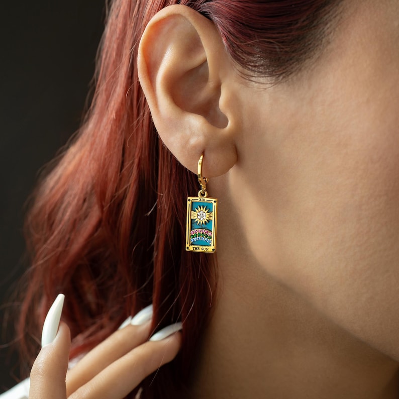 May include: Gold hoop earrings with a turquoise blue rectangular charm featuring a tarot card design with a sun and rainbow. The card reads "The Sun".