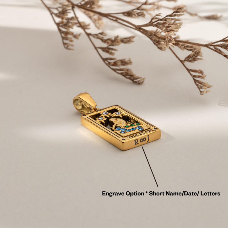 May include: Gold pendant necklace featuring a tarot card design with the words "The Star" and "Roo" on the front. The card is decorated with colorful gemstones.