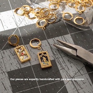 May include: Two gold earrings with a tarot card design. The earrings feature the "The Lovers" and "The Star" cards. The earrings are on a gray surface with gold jump rings.