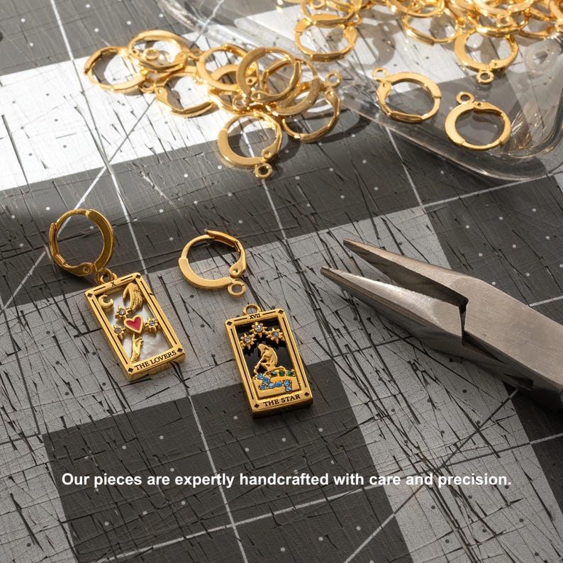 May include: Two gold earrings with a tarot card design. The earrings feature the "The Lovers" and "The Star" cards. The earrings are on a gray surface with gold jump rings.