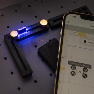 May include: A black metal L-shaped tool with two gold knobs and a blue light. The tool is on a gray surface with a grid pattern. A smartphone screen shows a measurement of 14.14mm x 3.85mm.