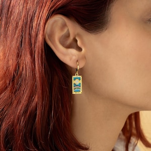 May include: Gold hoop earrings with a turquoise and gold tarot card charm featuring a sun and moon design. The card reads "The Sun".