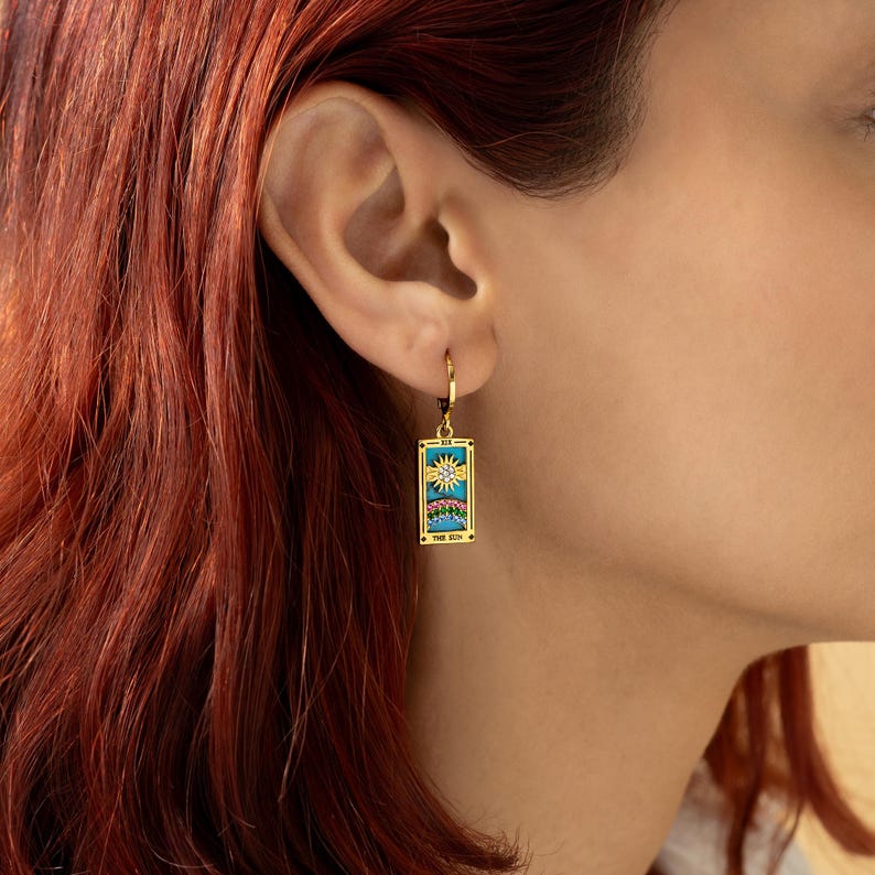 May include: Gold hoop earrings with a turquoise and gold tarot card charm featuring a sun and moon design. The card reads "The Sun".