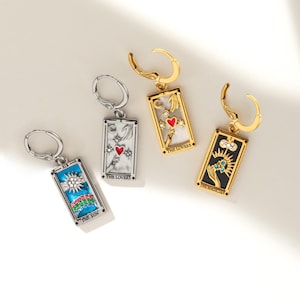 May include: Four silver and gold hoop earrings with tarot card charms. The charms feature colorful enamel and rhinestones. The cards are "The Lovers", "The Magician", and "The Sun".