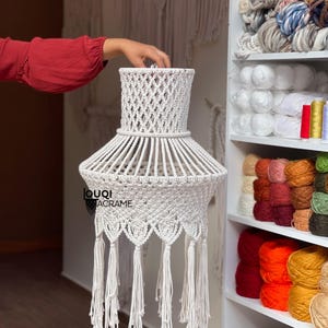 May include: White macrame lampshade with a fringe border. The lampshade is hanging from the ceiling and is in a room with a shelf full of yarn.