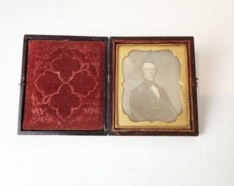Puede incluir: Una fotografía antigua de un hombre con traje oscuro y corbata, enmarcada en un marco dorado con un fondo de terciopelo rojo. La fotografía está en una caja de madera.