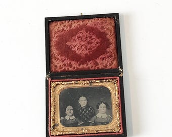 Puede incluir: Una fotografía antigua enmarcada en negro y dorado de tres personas. La foto está en una caja de madera forrada de terciopelo.