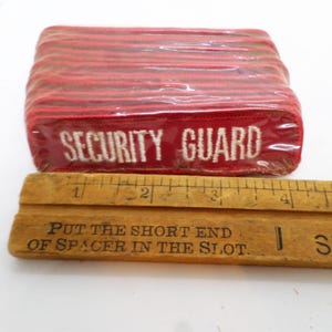 May include: A stack of red fabric patches with white embroidery that reads "SECURITY GUARD". The patches are approximately 3 inches long and are packaged in clear plastic.