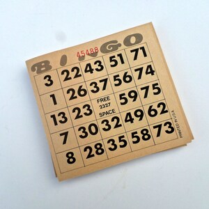 Puede incluir: Un cart&oacute;n de bingo marr&oacute;n con n&uacute;meros negros y la palabra "Bingo" impresa en letras rojas. El cart&oacute;n tiene un espacio libre en el centro y los n&uacute;meros 3, 22, 43, 51, 71, 1, 26, 37, 56, 74, 13, 23, 59, 75, 7, 30, 32, 49, 62, 8, 28, 35, 58, 73.