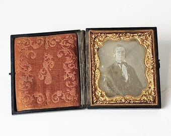 May include: A black velvet-lined wooden case with a gold-trimmed frame containing a black and white photograph of a man in a suit and tie.