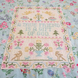 May include: A rectangular off-white fabric sampler featuring a cross-stitch design with the text A TRUE FRIEND IS A GIFT OF GOD. The embroidery includes yellow angels, pink and green floral motifs, small pine trees, and birds in a decorative border.