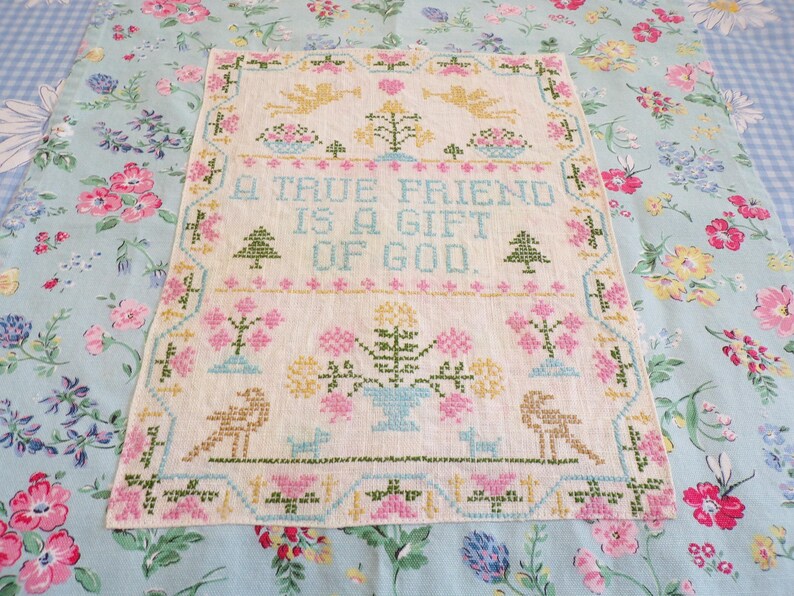 May include: A rectangular off-white fabric sampler featuring a cross-stitch design with the text A TRUE FRIEND IS A GIFT OF GOD. The embroidery includes yellow angels, pink and green floral motifs, small pine trees, and birds in a decorative border.