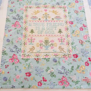 May include: A light blue fabric tea towel featuring a floral pattern with pink, yellow, and blue flowers. Centered on the towel is a rectangular cross-stitch sampler with the text A TRUE FRIEND IS A GIFT OF GOD, surrounded by birds, trees, and flowers.