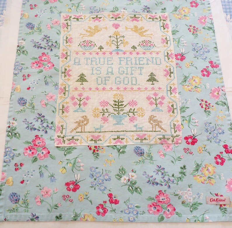 May include: A light blue fabric tea towel featuring a floral pattern with pink, yellow, and blue flowers. Centered on the towel is a rectangular cross-stitch sampler with the text A TRUE FRIEND IS A GIFT OF GOD, surrounded by birds, trees, and flowers.