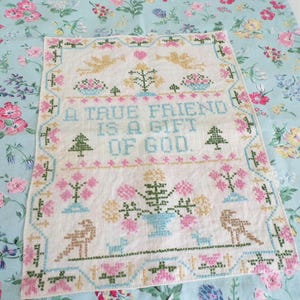 May include: A rectangular white fabric sampler featuring cross-stitch embroidery with the text A TRUE FRIEND IS A GIFT OF GOD. The design includes golden angels, pink and blue floral motifs, green trees, and birds, all framed by a decorative border.