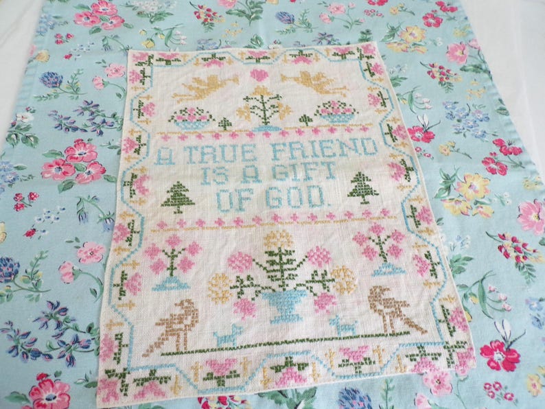 May include: A rectangular white fabric sampler featuring cross-stitch embroidery with the text A TRUE FRIEND IS A GIFT OF GOD. The design includes golden angels, pink and blue floral motifs, green trees, and birds, all framed by a decorative border.