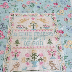 May include: A rectangular cross-stitch sampler on off-white fabric featuring the text A TRUE FRIEND IS A GIFT OF GOD, surrounded by embroidered motifs of angels, trees, and flowers, layered over a light blue fabric with a floral pattern.