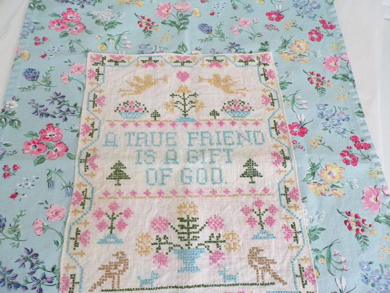 May include: A rectangular cross-stitch sampler on off-white fabric featuring the text A TRUE FRIEND IS A GIFT OF GOD, surrounded by embroidered motifs of angels, trees, and flowers, layered over a light blue fabric with a floral pattern.