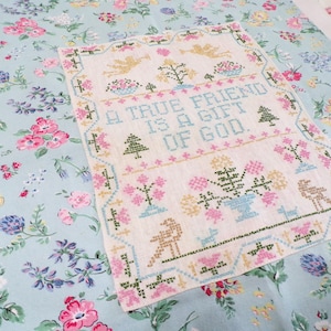 May include: A rectangular cross-stitch sampler on light beige fabric featuring the text A TRUE FRIEND IS A GIFT OF GOD in blue, surrounded by embroidered motifs of angels, trees, and flowers, centered on a light blue floral patterned fabric.