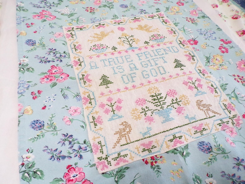 May include: A rectangular cross-stitch sampler on light beige fabric featuring the text A TRUE FRIEND IS A GIFT OF GOD in blue, surrounded by embroidered motifs of angels, trees, and flowers, centered on a light blue floral patterned fabric.