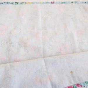 May include: A rectangular fabric piece featuring a light blue background with a soft floral pattern in shades of pink, yellow, and green. The edges are finished with a blue and white striped border. A small white fabric tag is attached at the corner.