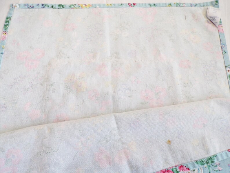 May include: A rectangular fabric piece featuring a light blue background with a soft floral pattern in shades of pink, yellow, and green. The edges are finished with a blue and white striped border. A small white fabric tag is attached at the corner.
