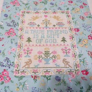 May include: A rectangular cross-stitch sampler on light fabric featuring the text A TRUE FRIEND IS A GIFT OF GOD in blue, surrounded by embroidered motifs of angels, flowers, trees, and birds in pink, green, yellow, and blue thread colors.