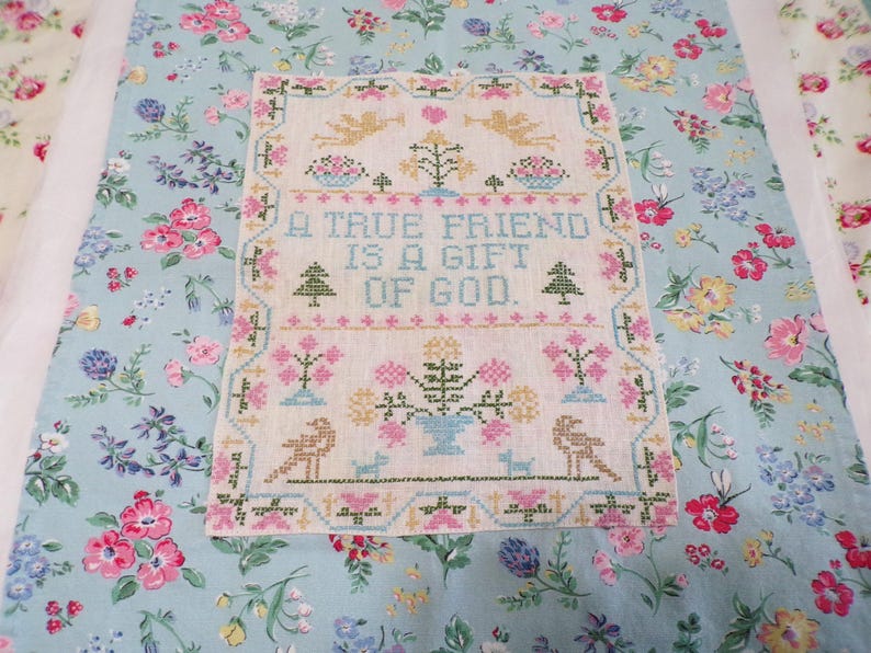 May include: A rectangular cross-stitch sampler on light fabric featuring the text A TRUE FRIEND IS A GIFT OF GOD in blue, surrounded by embroidered motifs of angels, flowers, trees, and birds in pink, green, yellow, and blue thread colors.