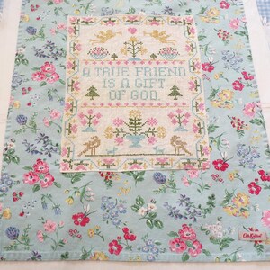 May include: A light blue fabric tea towel featuring a colorful floral pattern and a rectangular cross-stitch sampler in the center. The sampler displays the text A TRUE FRIEND IS A GIFT OF GOD, surrounded by embroidered birds, flowers, and trees.