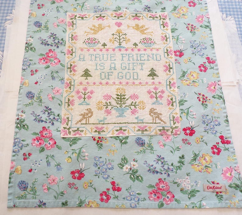 May include: A light blue fabric tea towel featuring a colorful floral pattern and a rectangular cross-stitch sampler in the center. The sampler displays the text A TRUE FRIEND IS A GIFT OF GOD, surrounded by embroidered birds, flowers, and trees.