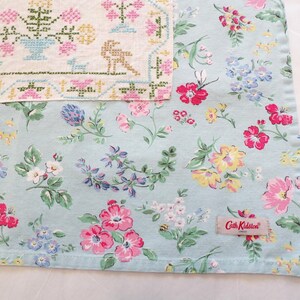 May include: A light blue fabric featuring a colorful floral pattern with pink, yellow, and blue flowers, layered beneath a cream-colored textile with a cross-stitched design of flowers and animals. A Cath Kidston label is sewn onto the fabric.