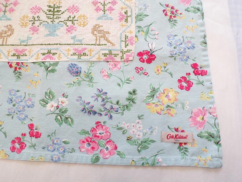 May include: A light blue fabric featuring a colorful floral pattern with pink, yellow, and blue flowers, layered beneath a cream-colored textile with a cross-stitched design of flowers and animals. A Cath Kidston label is sewn onto the fabric.