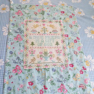 May include: A rectangular tea towel featuring a light blue background with a floral pattern of pink, yellow, and white flowers. Centered is a white fabric panel with cross-stitched text reading A TRUE FRIEND IS A GIFT OF GOD, surrounded by motifs of angels, trees, birds, and flowers.