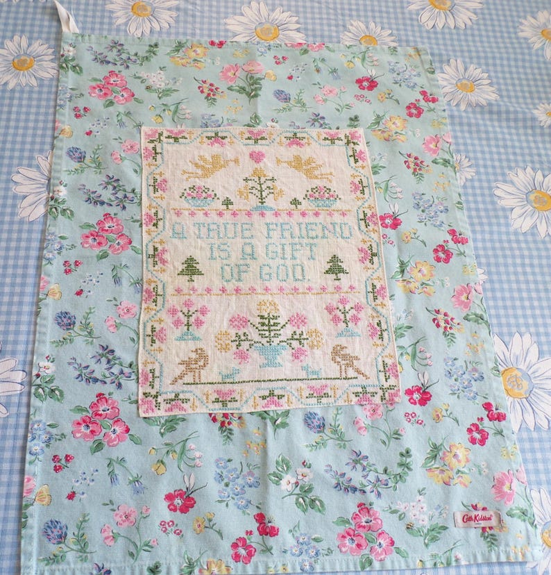 May include: A rectangular tea towel featuring a light blue background with a floral pattern of pink, yellow, and white flowers. Centered is a white fabric panel with cross-stitched text reading A TRUE FRIEND IS A GIFT OF GOD, surrounded by motifs of angels, trees, birds, and flowers.