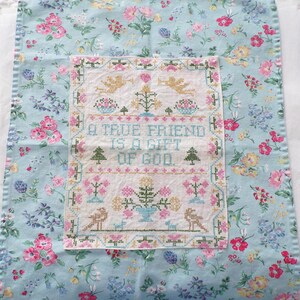 May include: A rectangular fabric panel featuring a central white cross-stitch embroidery on a light blue floral background. The embroidery displays the text A TRUE FRIEND IS A GIFT OF GOD, surrounded by motifs of angels, trees, and flowers.
