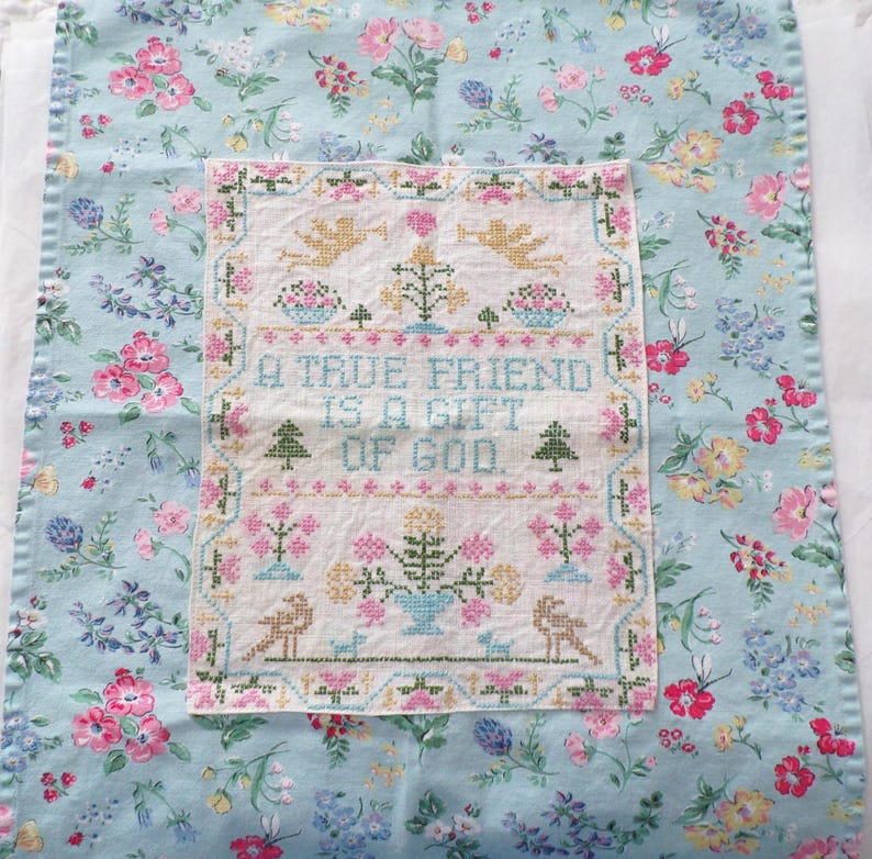 May include: A rectangular fabric panel featuring a central white cross-stitch embroidery on a light blue floral background. The embroidery displays the text A TRUE FRIEND IS A GIFT OF GOD, surrounded by motifs of angels, trees, and flowers.