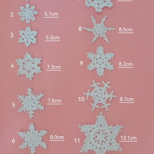 Könnte beinhalten: Elf weiße, gehäkelte Schneeflocken-Ornamente in verschiedenen Größen, von 3,2 cm bis 13,1 cm Durchmesser, werden vor einem rosa Hintergrund gezeigt. Jede Schneeflocke hat ein einzigartiges Design, mit Messungen in Zentimetern.