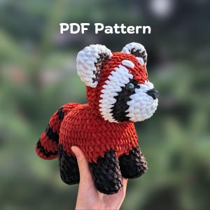 May include: A crocheted red panda toy with a fluffy texture, featuring red, black, and white colours. The text "PDF Pattern" is visible. The toy is held in a hand. The red panda has a black nose and eyes.