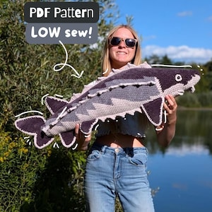 May include: A large, handmade crochet sturgeon fish plush toy featuring a dark charcoal and light gray color scheme with textured fins and a white eye. The image includes text that reads PDF Pattern LOW sew! with a white arrow pointing to the fish.