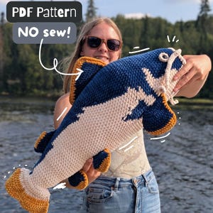 Peut inclure: Un poisson-chat en crochet avec un dos bleu foncé, un ventre crème et des nageoires ainsi qu'une queue jaune moutarde. Le texte sur l'image indique PDF Pattern NO sew! Le poisson possède des moustaches blanches et des yeux sombres en laine.