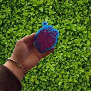 May include: A handmade, felt ornament in shades of blue and red. The ornament features a heart and floral design, with a grid pattern on the red portion. The ornament is held against a backdrop of green foliage.