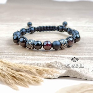 Può includere: Braccialetto di perline nere con chiusura macramè. Il braccialetto presenta un mix di perline nere opache e lucide, perline di strass argentate e perline rosso scuro. Il braccialetto è fatto a mano e il logo "Crystal Crafted Handmade" è visibile.