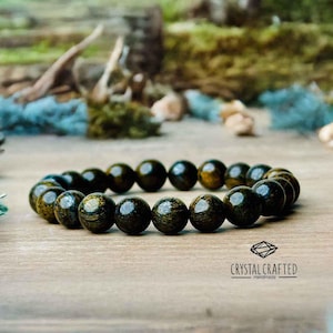 Puede incluir: Un brazalete de cuentas de madera de color verde oscuro y dorado. El brazalete está hecho a mano y tiene un aspecto natural y rústico.