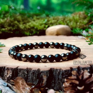 Pode incluir: Uma pulseira de contas pretas com contas foscas e metálicas alternadas. A pulseira é exibida em uma superfície de madeira com um fundo natural. A marca "Crystal Crafted Handmade" é visível.