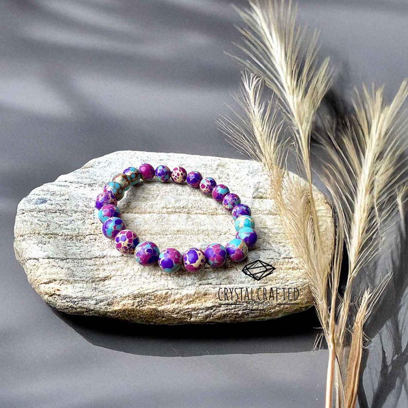 A handcrafted Sea Sediment Jasper Bracelet with vibrant purple, blue, and earthy tones, resting on a natural stone slab. A calming accessory for grounding and balance.