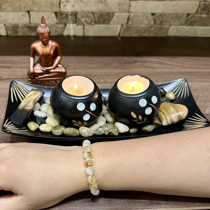 K&ouml;nnte beinhalten: Ein schwarzes Dekotablett mit zwei brennenden Kerzen in schwarzen Haltern, verziert mit wei&szlig;en Punkten und Blattmustern. Eine kleine Buddha-Statue aus Bronze ist im Hintergrund zu sehen. Ein Armband mit gelben und wei&szlig;en Perlen ist sichtbar.